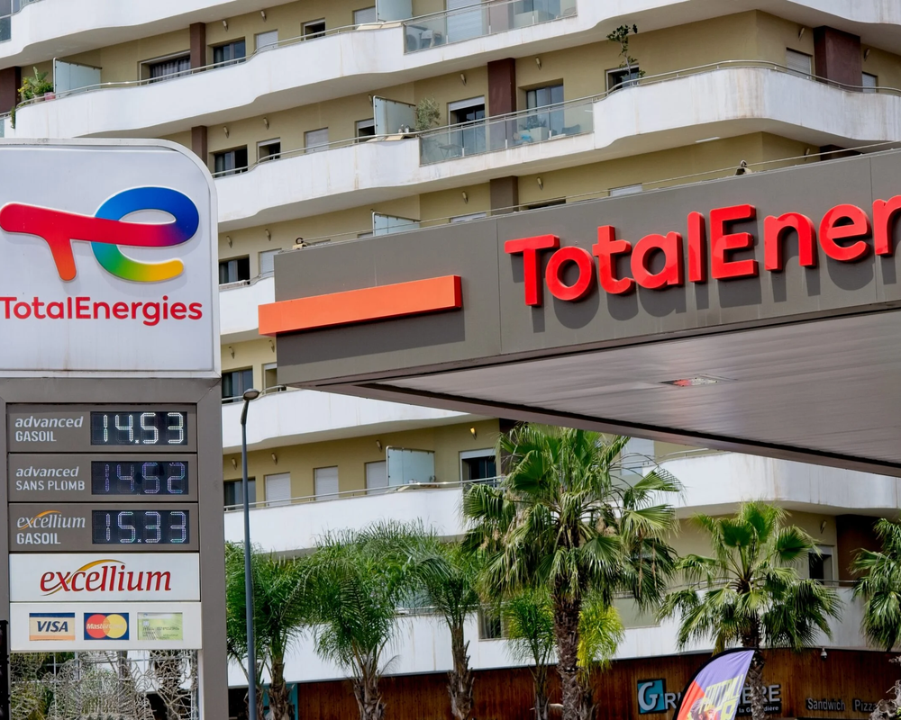 TotalEnergies gas station with price display showing fuel costs (gasoil at 14.53, sans plumb at 14.52, excellium gasoil at 15.93), located in front of a modern apartment building with palm trees in the foreground.