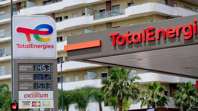 TotalEnergies gas station with price display showing fuel costs (gasoil at 14.53, sans plumb at 14.52, excellium gasoil at 15.93), located in front of a modern apartment building with palm trees in the foreground.