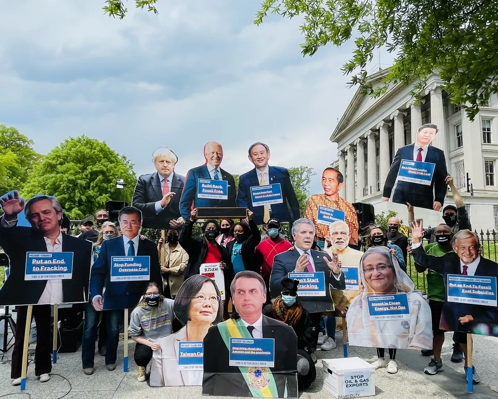Protesters gather in front of a classical white building with columns, holding oversized cutouts of world leaders' faces and blue signs demanding climate action. Trees frame the scene under a cloudy sky.