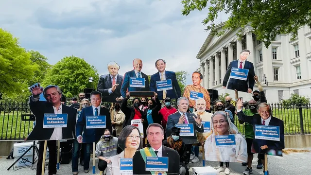 Protesters gather in front of a classical white building with columns, holding oversized cutouts of world leaders' faces and blue signs demanding climate action. Trees frame the scene under a cloudy sky.