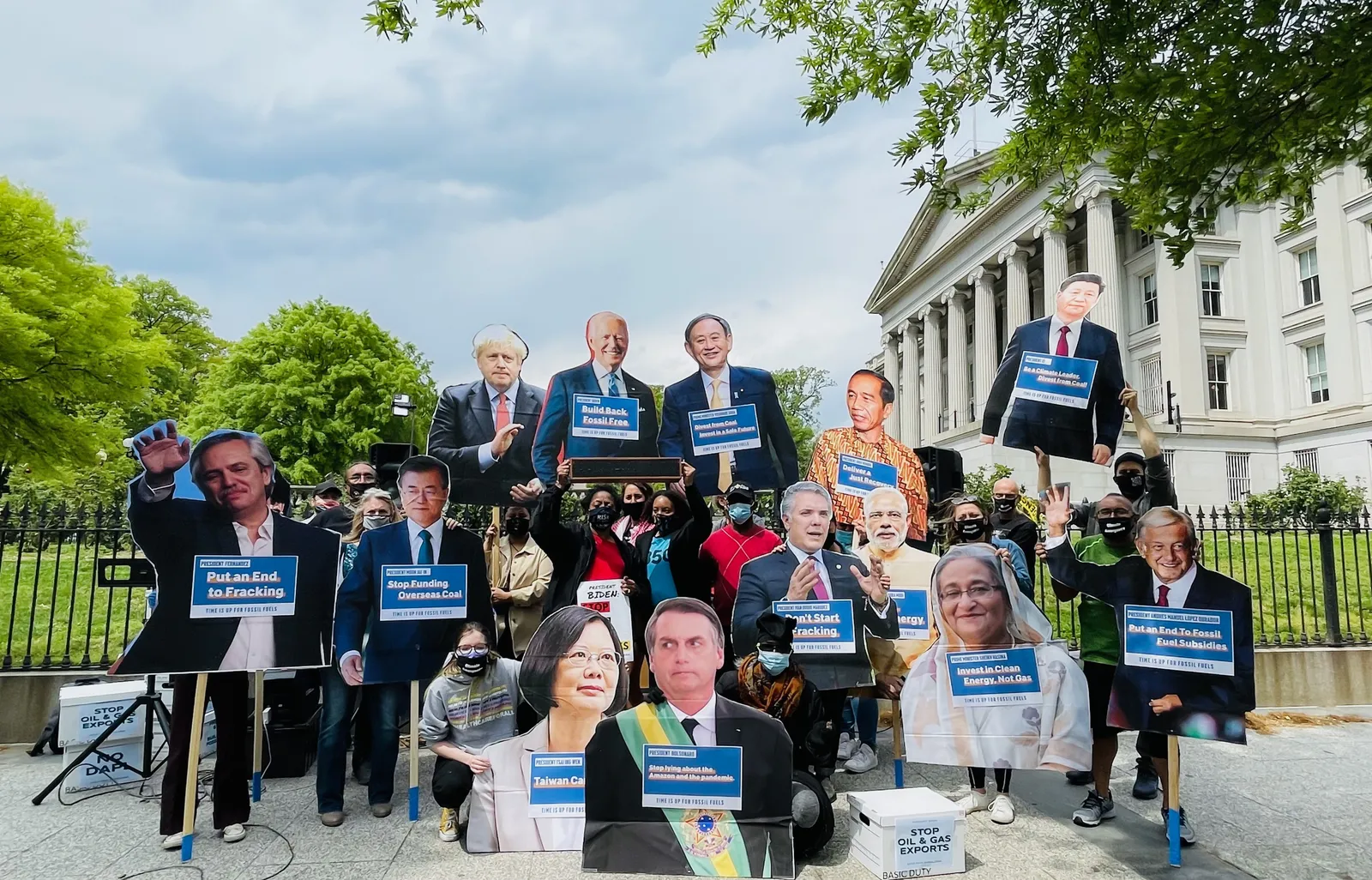 Protesters gather in front of a classical white building with columns, holding oversized cutouts of world leaders' faces and blue signs demanding climate action. Trees frame the scene under a cloudy sky.