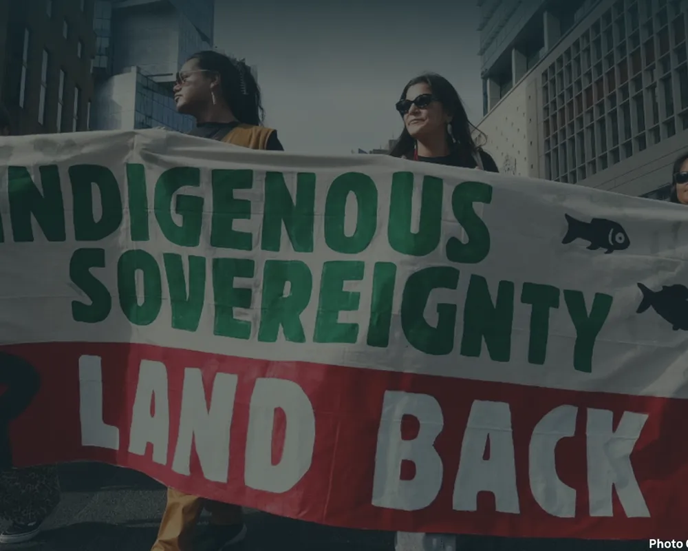 Protesters hold a banner reading "Indigenous Sovereignty Land Back" during a demonstration in an urban setting.
