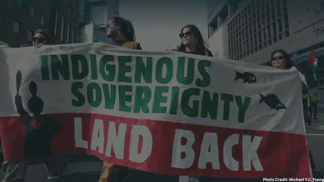 Protesters hold a banner reading "Indigenous Sovereignty Land Back" during a demonstration in an urban setting.