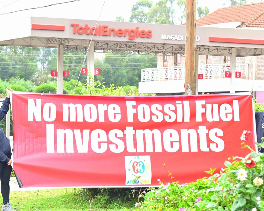 Aktivist*innen halten einen Banner auf dem "No more Fossil Fuel Investments" steht. Im Hintergrund ist eine Tankstelle von TOTAL zu sehen.