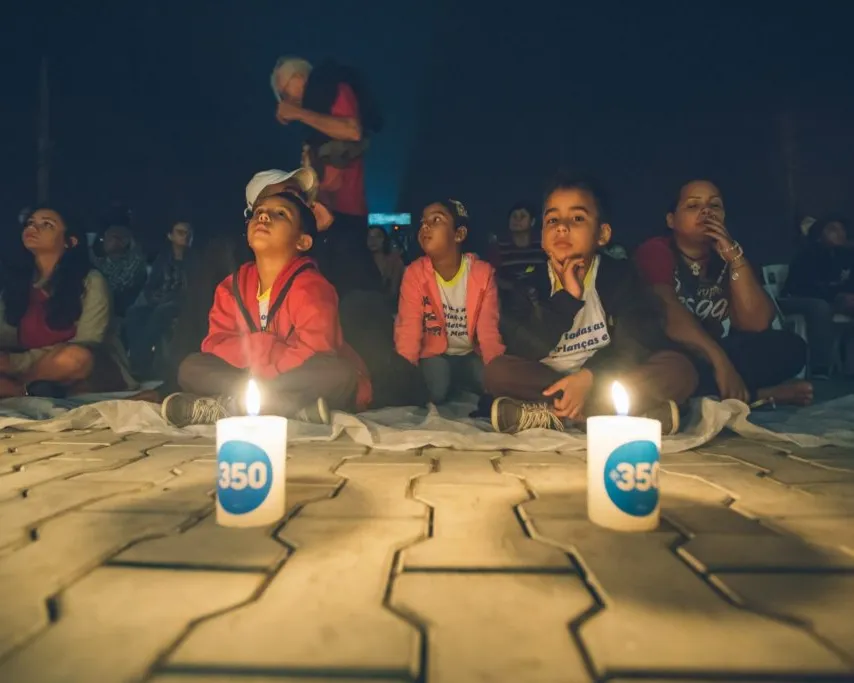 Children sit on the ground watching a performance, with two lit candles bearing "350" logos in the foreground. A performer is visible in the background against a dark setting.