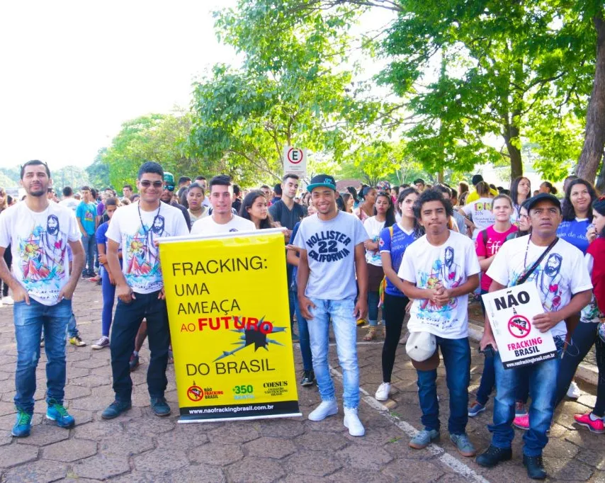 Protesters gather outdoors holding yellow and white signs against fracking in Brazil. Large crowd stands under green trees in sunny park setting, demonstrating environmental concerns about Brazil's future.