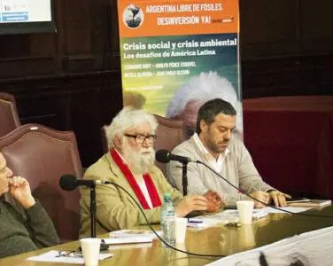 Three panelists at a conference table discussing social and environmental crisis in Latin America. Banner visible in background. Indoor setting with microphones and water bottles on table.