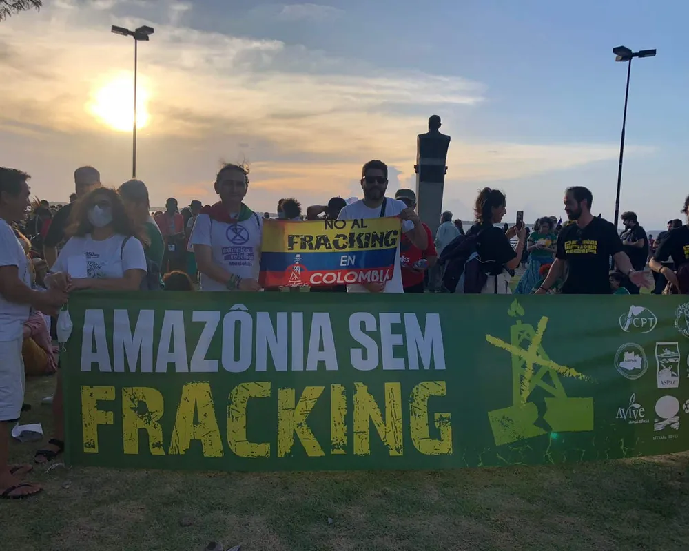 Environmental protesters gather at sunset holding banners against fracking in the Amazon and Colombia. The crowd displays signs reading "AMAZÔNIA SEM FRACKING" and "NO AL FRACKING EN COLOMBIA" under street lights.