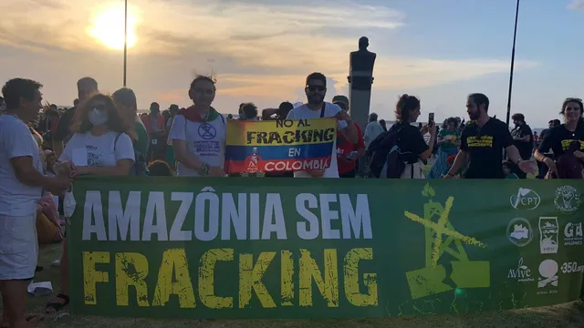 Environmental protesters gather at sunset holding banners against fracking in the Amazon and Colombia. The crowd displays signs reading "AMAZÔNIA SEM FRACKING" and "NO AL FRACKING EN COLOMBIA" under street lights.
