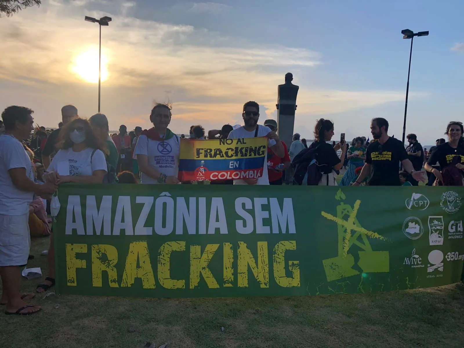 Environmental protesters gather at sunset holding banners against fracking in the Amazon and Colombia. The crowd displays signs reading "AMAZÔNIA SEM FRACKING" and "NO AL FRACKING EN COLOMBIA" under street lights.