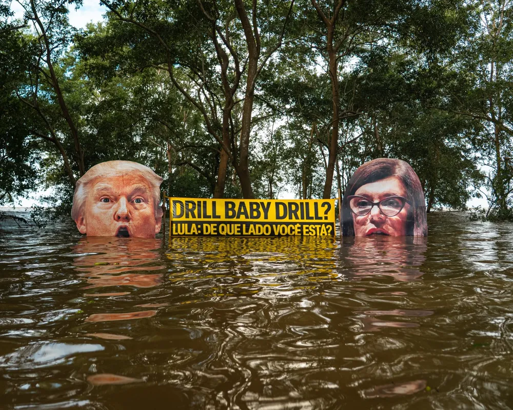 Political art installation showing two faces partially submerged in floodwater with Portuguese text on yellow sign between them, surrounded by trees, highlighting climate change concerns.