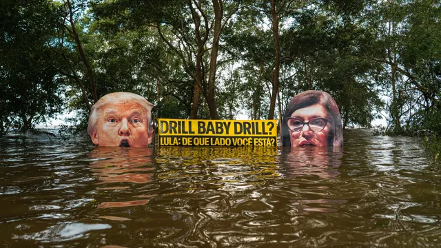 Political art installation showing two faces partially submerged in floodwater with Portuguese text on yellow sign between them, surrounded by trees, highlighting climate change concerns.