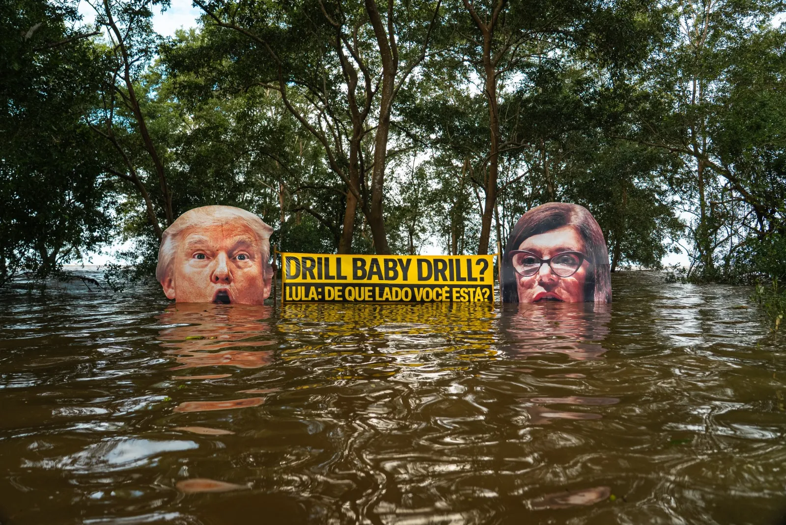 Political art installation showing two faces partially submerged in floodwater with Portuguese text on yellow sign between them, surrounded by trees, highlighting climate change concerns.
