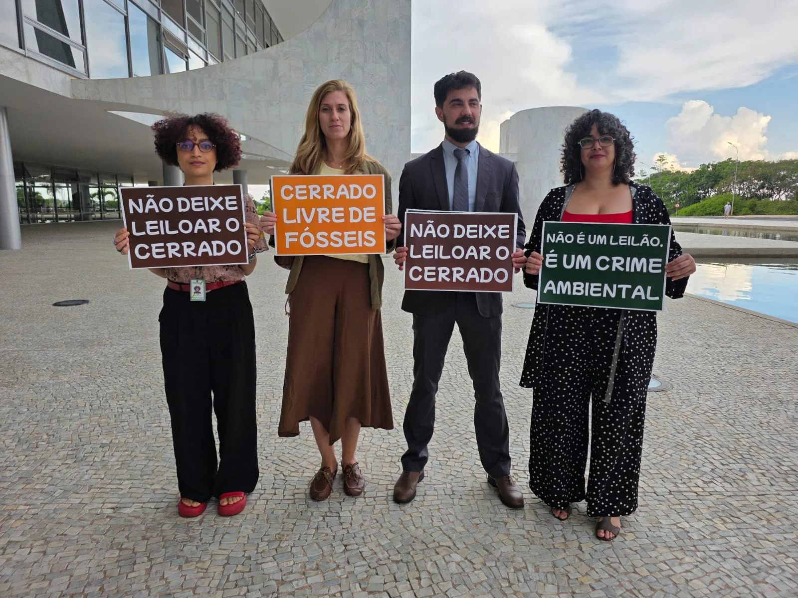 Four activists stand in front of a modern government building in Brazil holding protest signs in Portuguese that read "Don't let the Cerrado be auctioned," "Cerrado Free of Fossils," and "It's not an auction, it's an environmental crime."