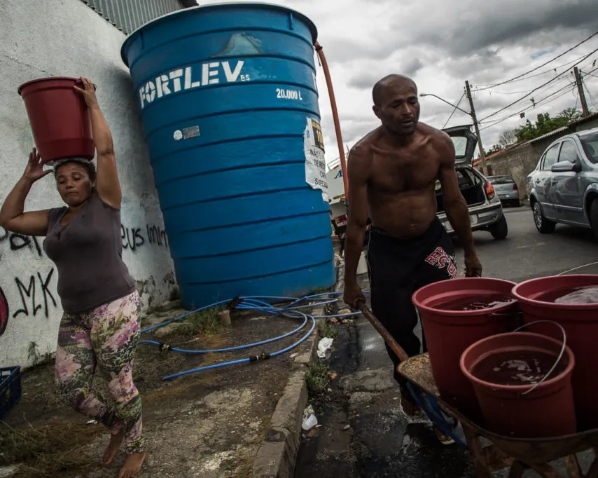 Two people collect water from a large blue Fortleva water tank on a street. A woman carries a red bucket on her head while a shirtless man stands beside red buckets on a cart under cloudy skies.