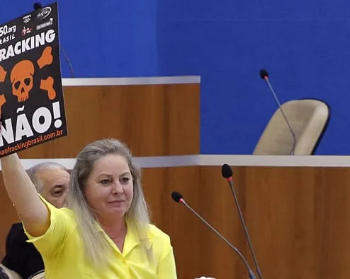 A woman in a yellow shirt holds up an anti-fracking protest sign with a skull symbol and "NÃO!" text during what appears to be a legislative or government meeting session.
