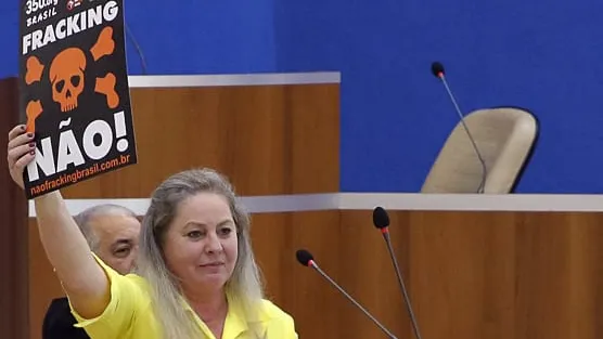 A woman in a yellow shirt holds up an anti-fracking protest sign with a skull symbol and "NÃO!" text during what appears to be a legislative or government meeting session.