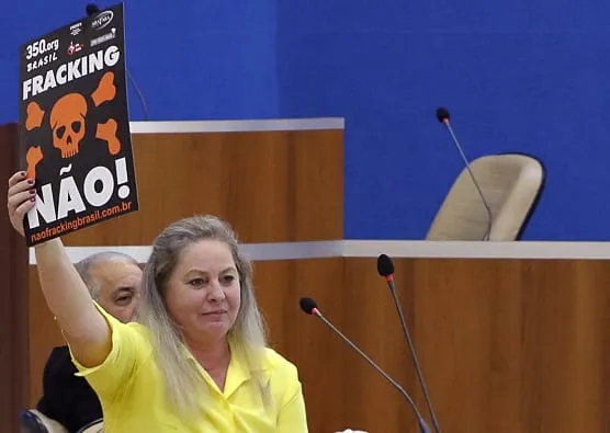 A woman in a yellow shirt holds up an anti-fracking protest sign with a skull symbol and "NÃO!" text during what appears to be a legislative or government meeting session.