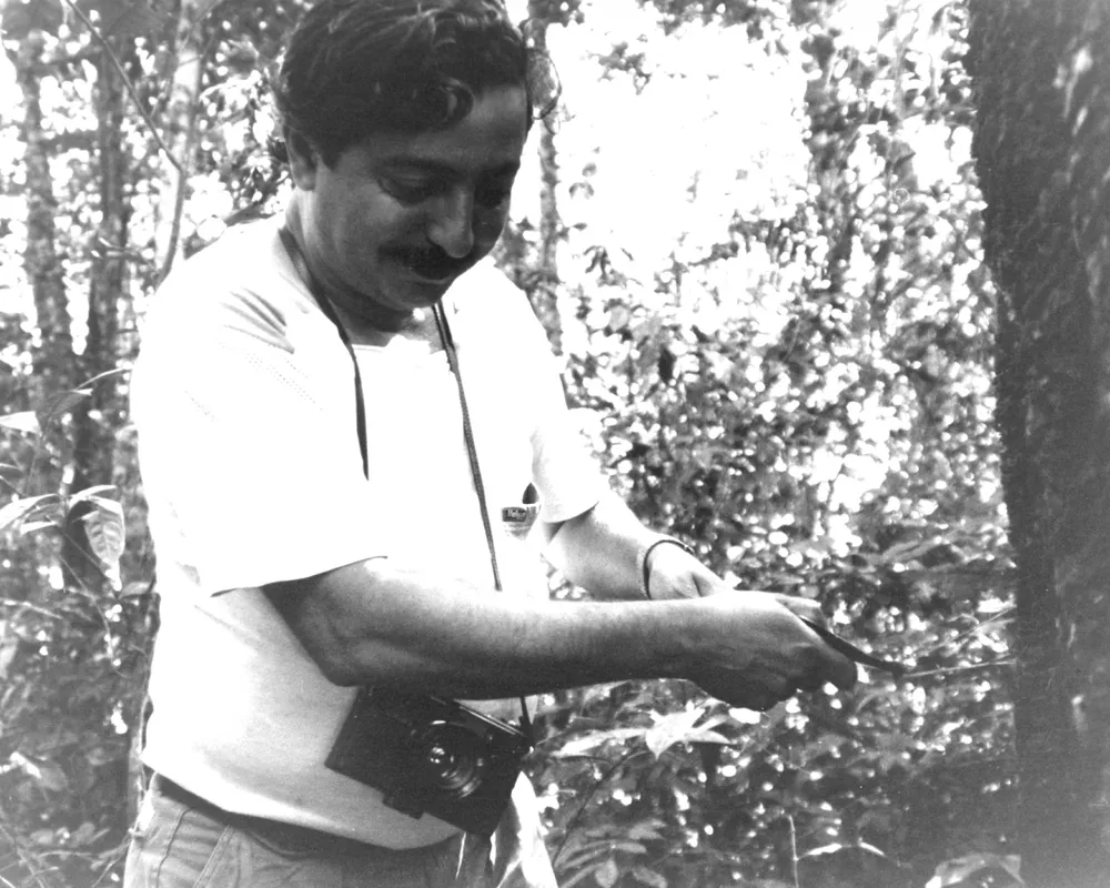 A man in a white short-sleeved shirt examines or works with something in his hands outdoors, surrounded by trees and foliage in this black and white photograph.