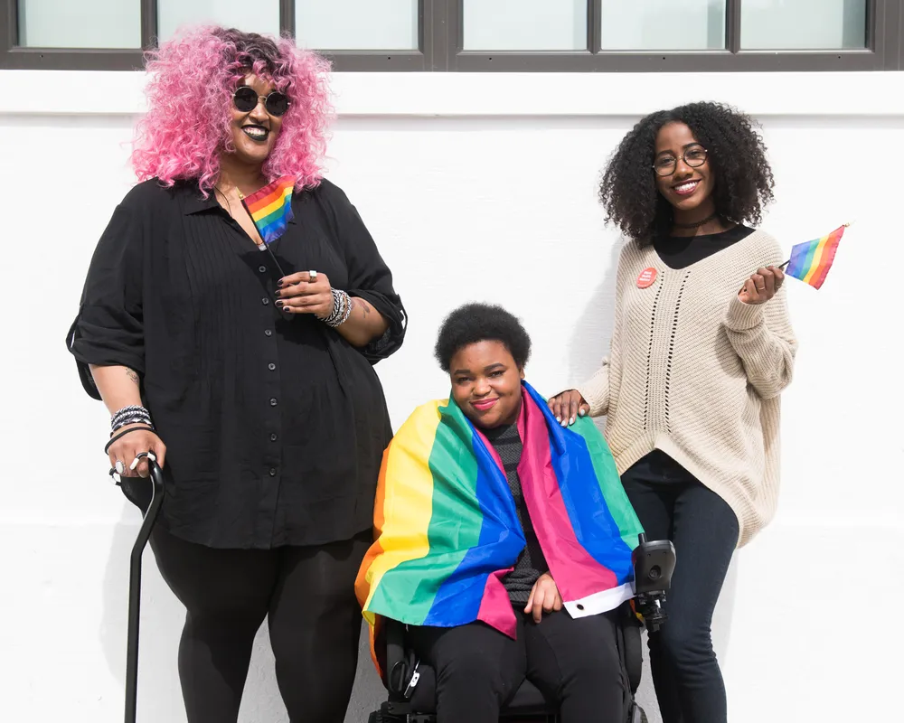 Three Black and disabled folx decked out happily in rainbow pride flags. On the left, a non-binary person holds a cane in one hand and waves a mini flag in the other. In the middle, a non-binary person sits in their power wheelchair, draped in a large fla