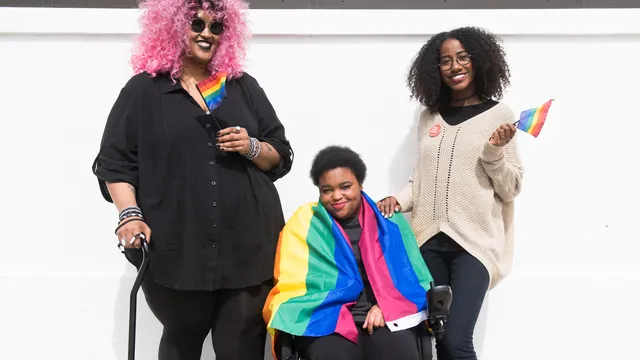 Three Black and disabled folx decked out happily in rainbow pride flags. On the left, a non-binary person holds a cane in one hand and waves a mini flag in the other. In the middle, a non-binary person sits in their power wheelchair, draped in a large fla