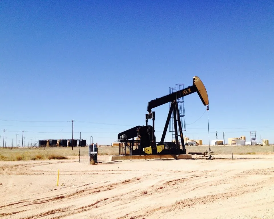 An oil pump jack operates in an arid desert landscape under a clear blue sky, surrounded by sandy terrain, utility poles, and industrial buildings in the background.