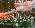 Protesters holding a banner with red smoke flares during an environmental demonstration. The banner appears to reference fossil gas and includes text about going nowhere, suggesting climate activism.