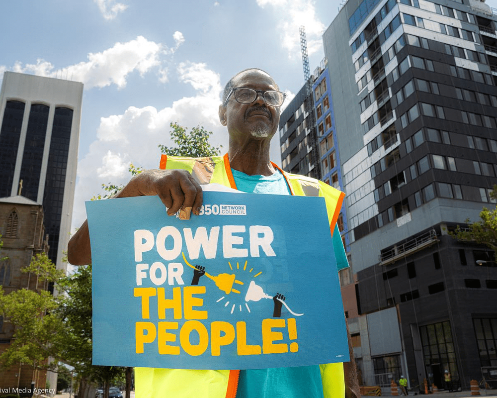 An activist wearing glasses and a colorful vest holds a blue sign reading "POWER FOR THE PEOPLE!" at a rally with modern buildings and cloudy skies in the background.