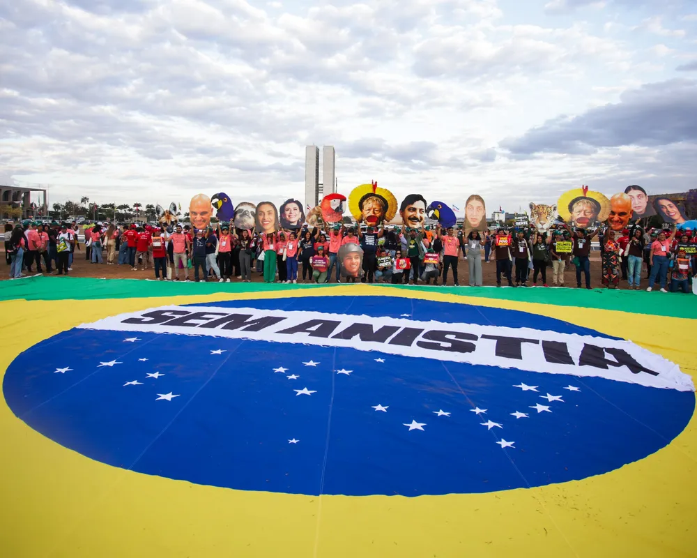 Protesters gather around a large Brazilian flag on the ground in Brasília, holding signs and wearing masks depicting political figures during a demonstration under cloudy skies.
