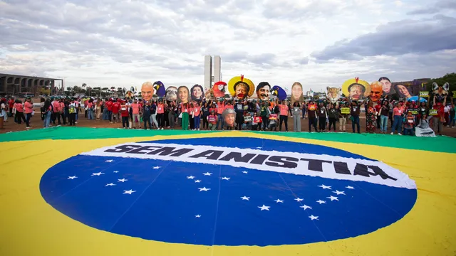 Protesters gather around a large Brazilian flag on the ground in Brasília, holding signs and wearing masks depicting political figures during a demonstration under cloudy skies.