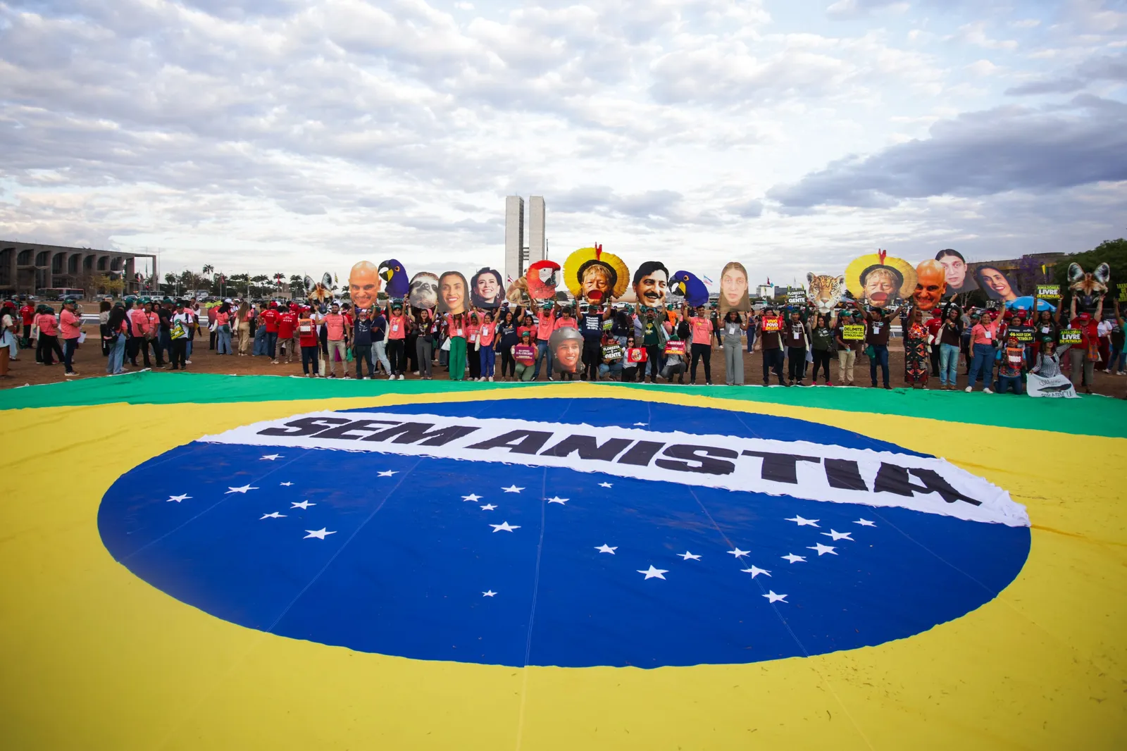 Protesters gather around a large Brazilian flag on the ground in Brasília, holding signs and wearing masks depicting political figures during a demonstration under cloudy skies.