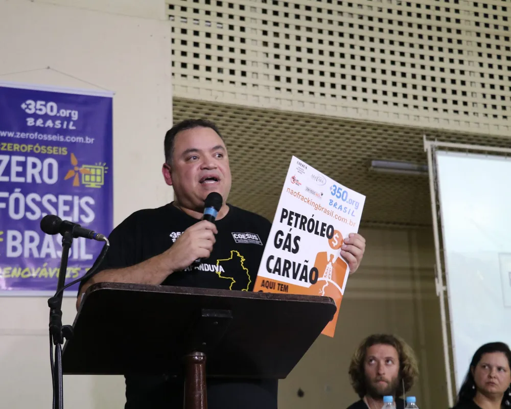 A speaker at a podium holds a sign about petroleum, gas, and coal at a Zero Fossil Fuels Brazil event. Purple banners display the 350.org campaign branding, with attendees seated behind the speaker.