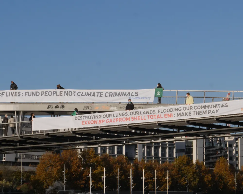 Climate activists display protest banners on a pedestrian bridge. Messages demand funding for people over climate action and call out fossil fuel companies. Modern buildings and autumn trees visible below.