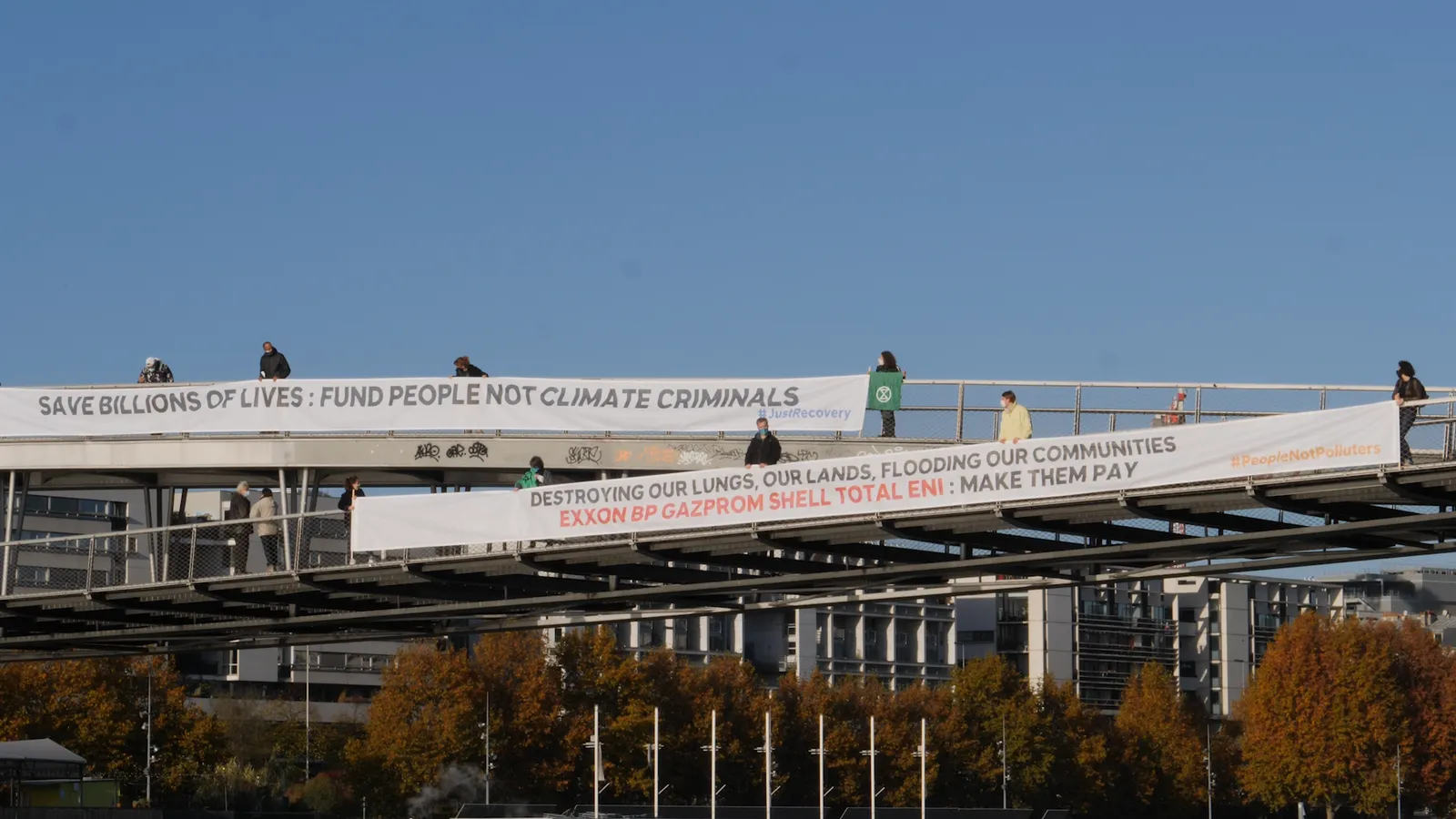 Climate activists display protest banners on a pedestrian bridge. Messages demand funding for people over climate action and call out fossil fuel companies. Modern buildings and autumn trees visible below.