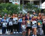 A large group of people gathered outdoors holding photos and signs, appearing to be at a memorial or awareness event. Trees and buildings are visible in the background. The crowd stands together in solidarity.