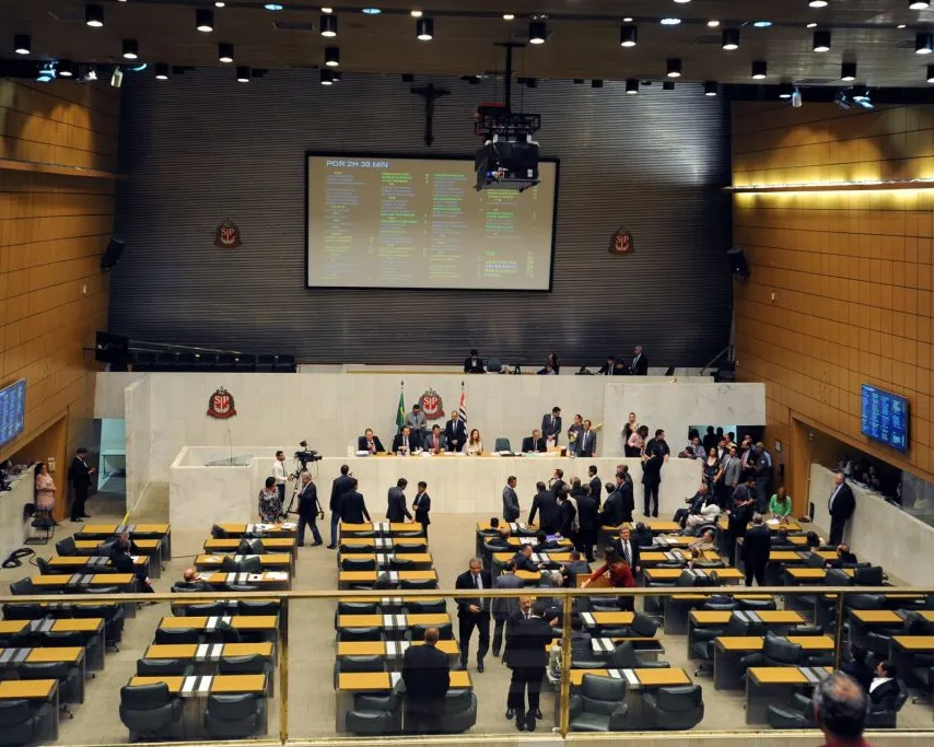 Modern legislative chamber with wooden accents and tiered seating. People gather on the floor while officials sit at the elevated podium. A large screen displays information above the assembly area.