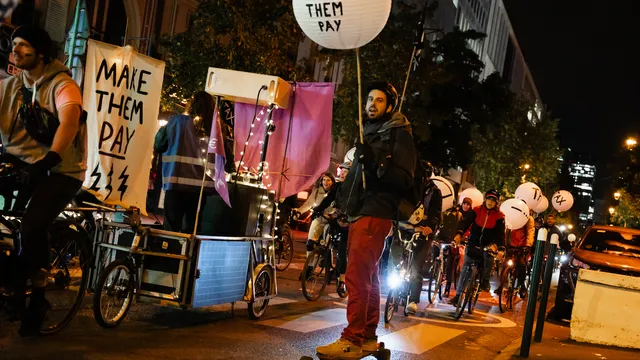 Night protest with cyclists holding "MAKE THEM PAY" signs, featuring illuminated banners and creative mobile displays on bikes gathering in an urban street demonstration.