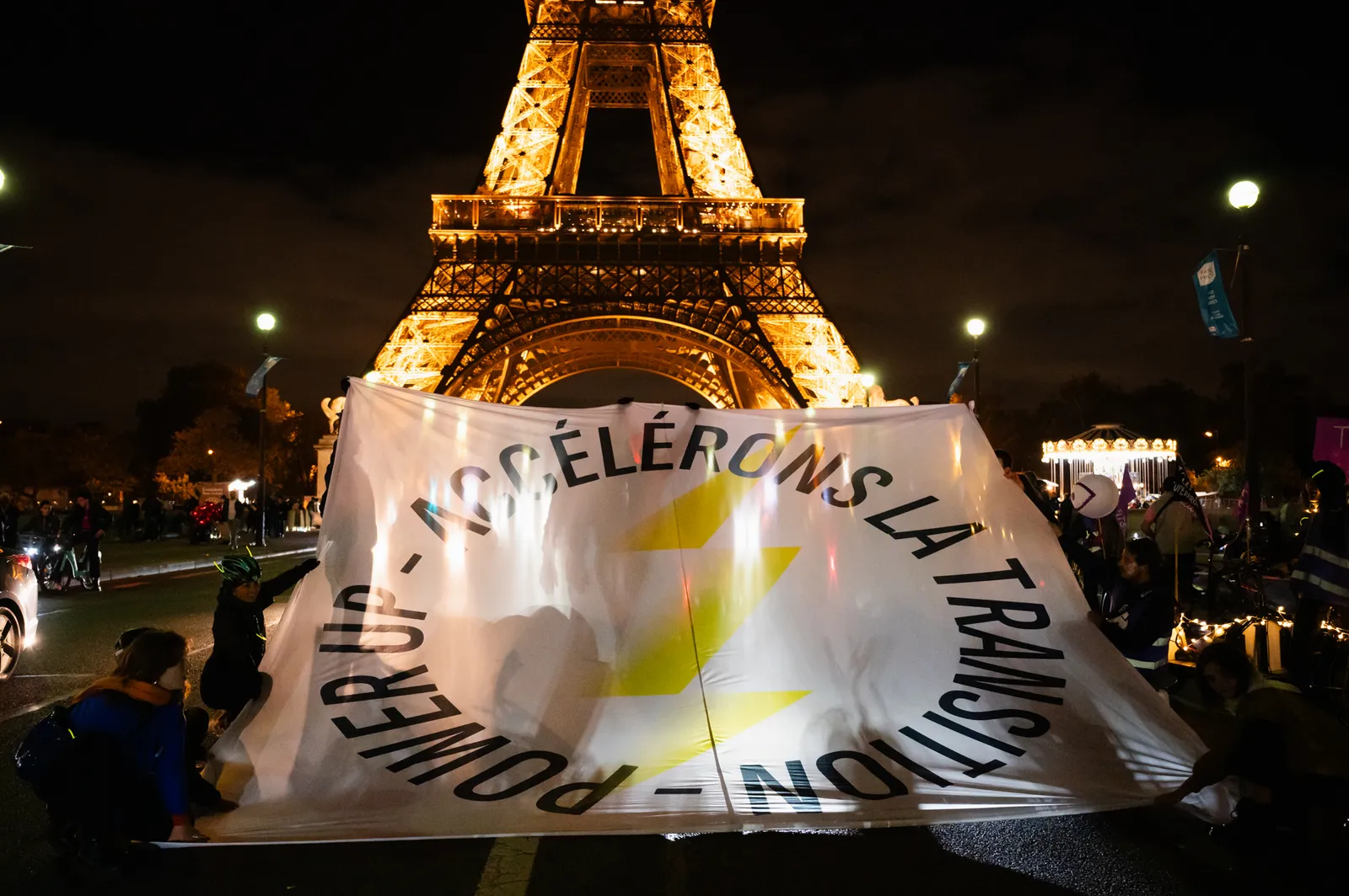 Protesters hold a large banner in front of the illuminated Eiffel Tower at night in Paris. The banner features French text and yellow lightning bolt symbols, with crowds gathered around during what appears to be a demonstration.