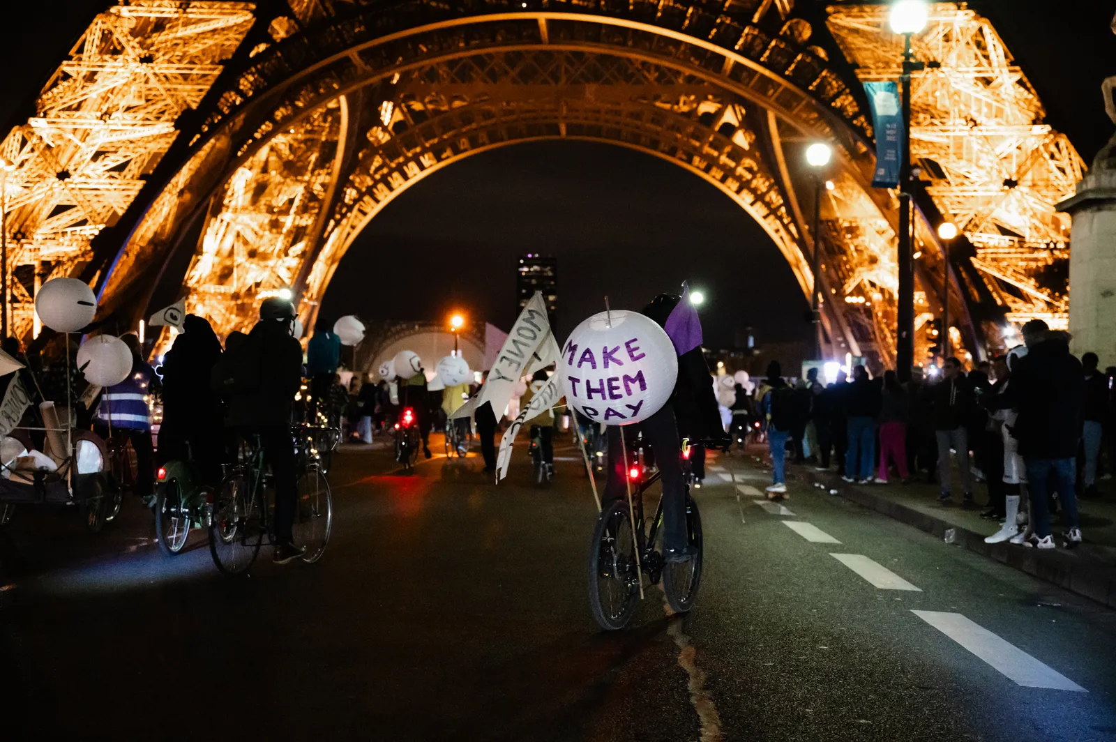 Night protest at the Eiffel Tower in Paris with cyclists holding illuminated signs reading "WAKE THEM" as crowds gather beneath the iconic landmark's golden arches.