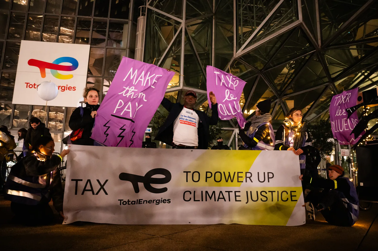Climate activists protest outside TotalEnergies headquarters holding signs demanding the company pay taxes and support climate justice. Banner reads "Tax TotalEnergies to power up climate justice."