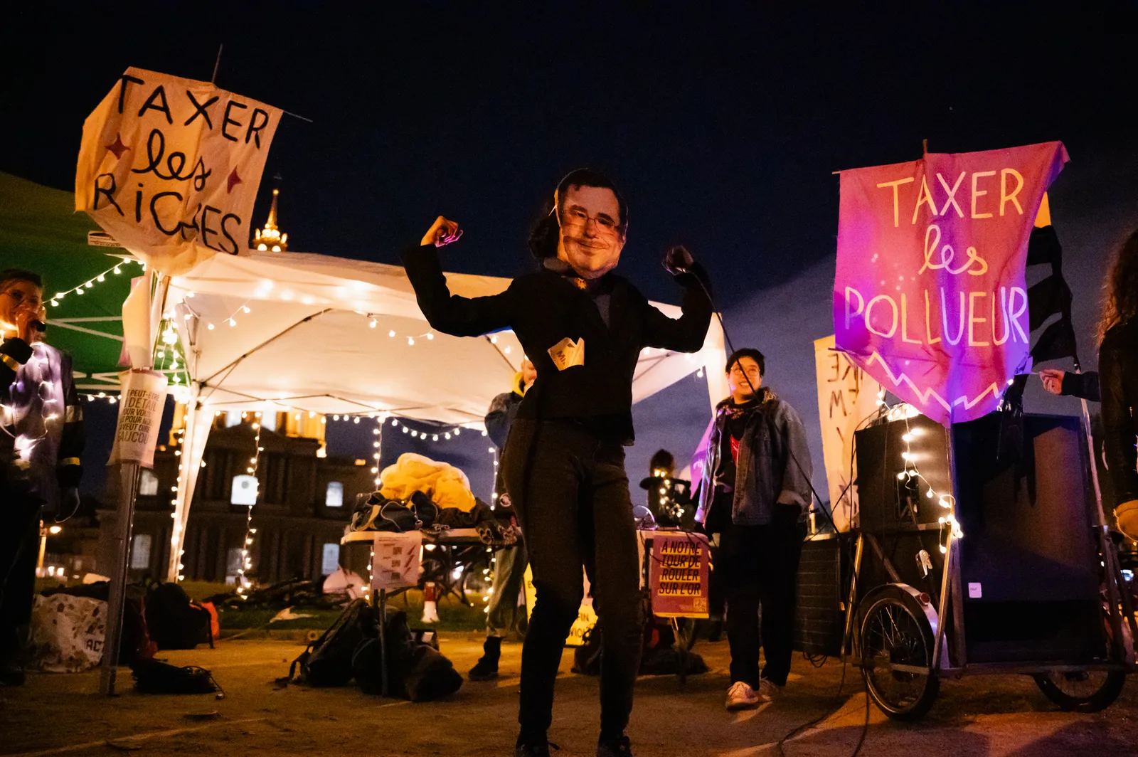 A nighttime protest with demonstrators holding illuminated signs reading "TAXER les RICHES" and "TAXER les POLLUEURS" in French, calling to tax the wealthy and polluters under string lights.