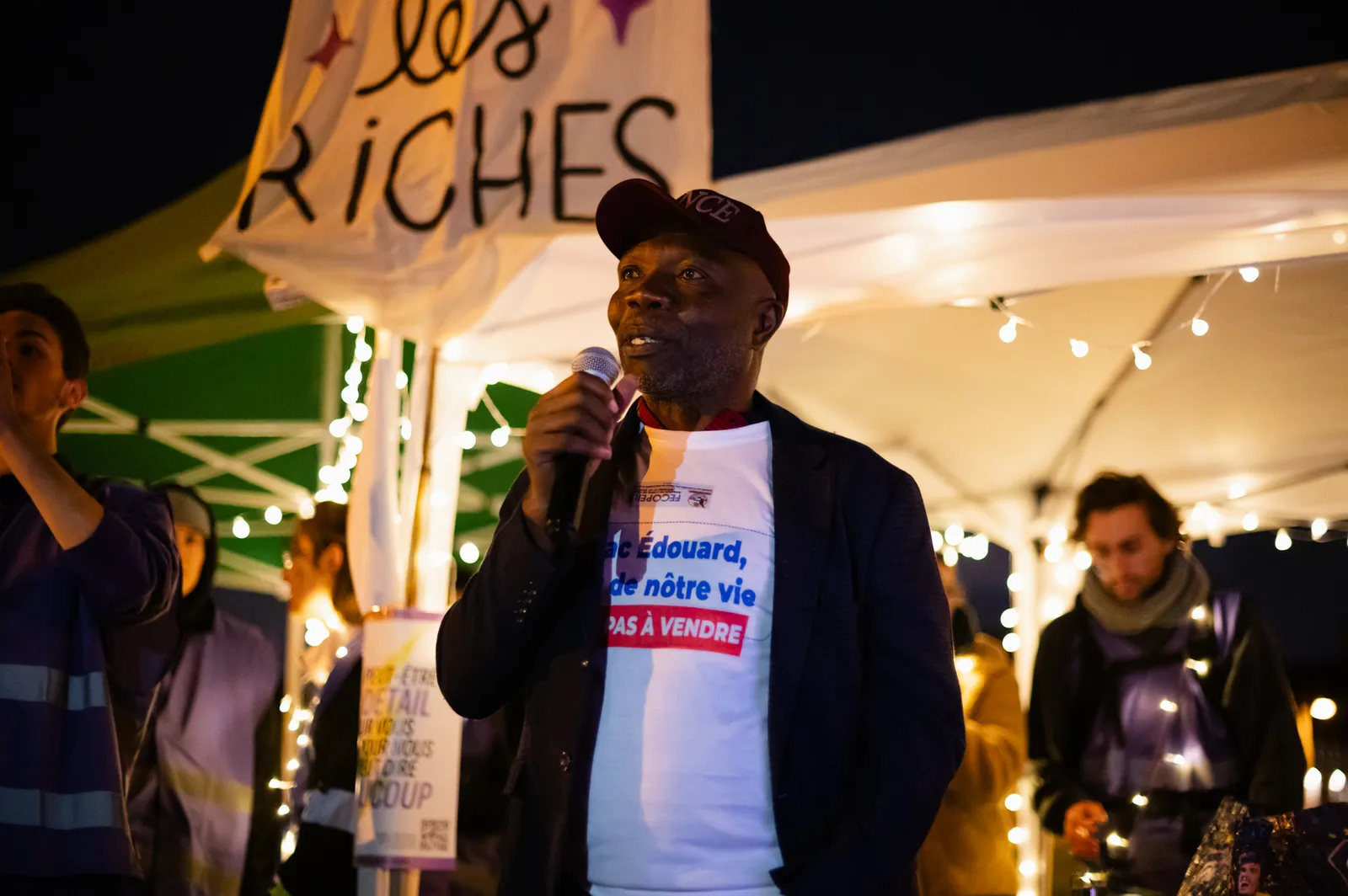A man in a cap speaks at a nighttime gathering under a tent with string lights. He wears a white shirt with French text. A banner reading "RICHES" is visible above, and people with candles stand nearby.