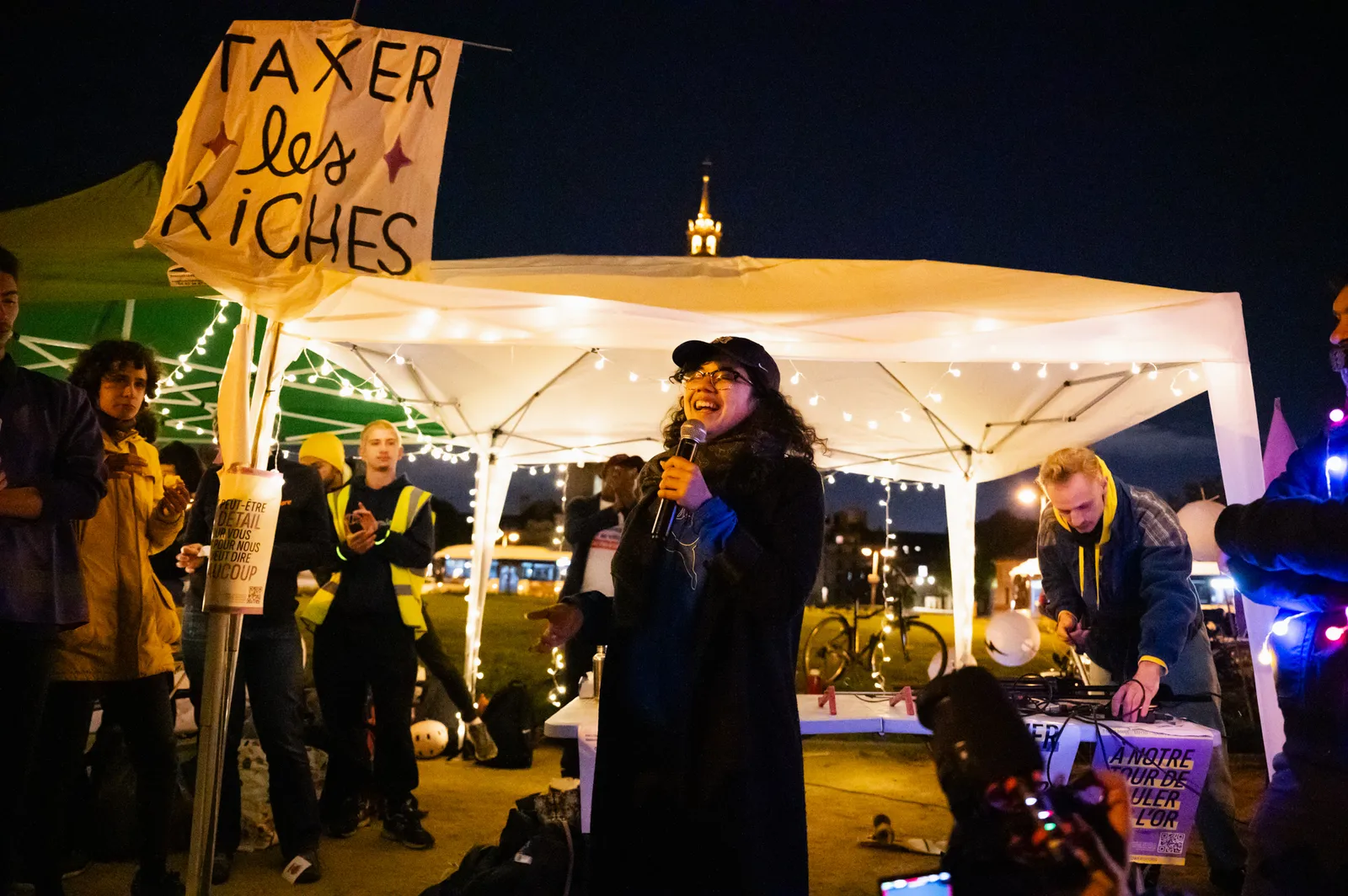 A nighttime protest scene with people gathered under a white tent. A woman speaks into a microphone while someone holds a sign reading "TAXER les RICHES" (Tax the Rich). City lights visible in background.