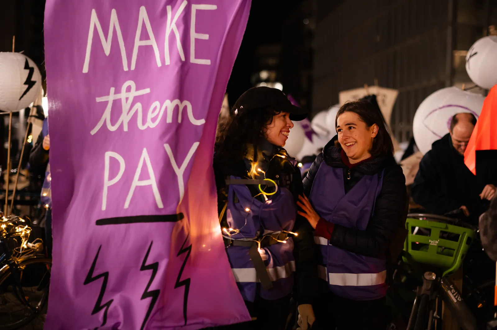 Two women smile beside a purple banner reading "MAKE them PAY" with lightning bolts at a nighttime gathering. People and props visible in background, suggesting a protest or community event.