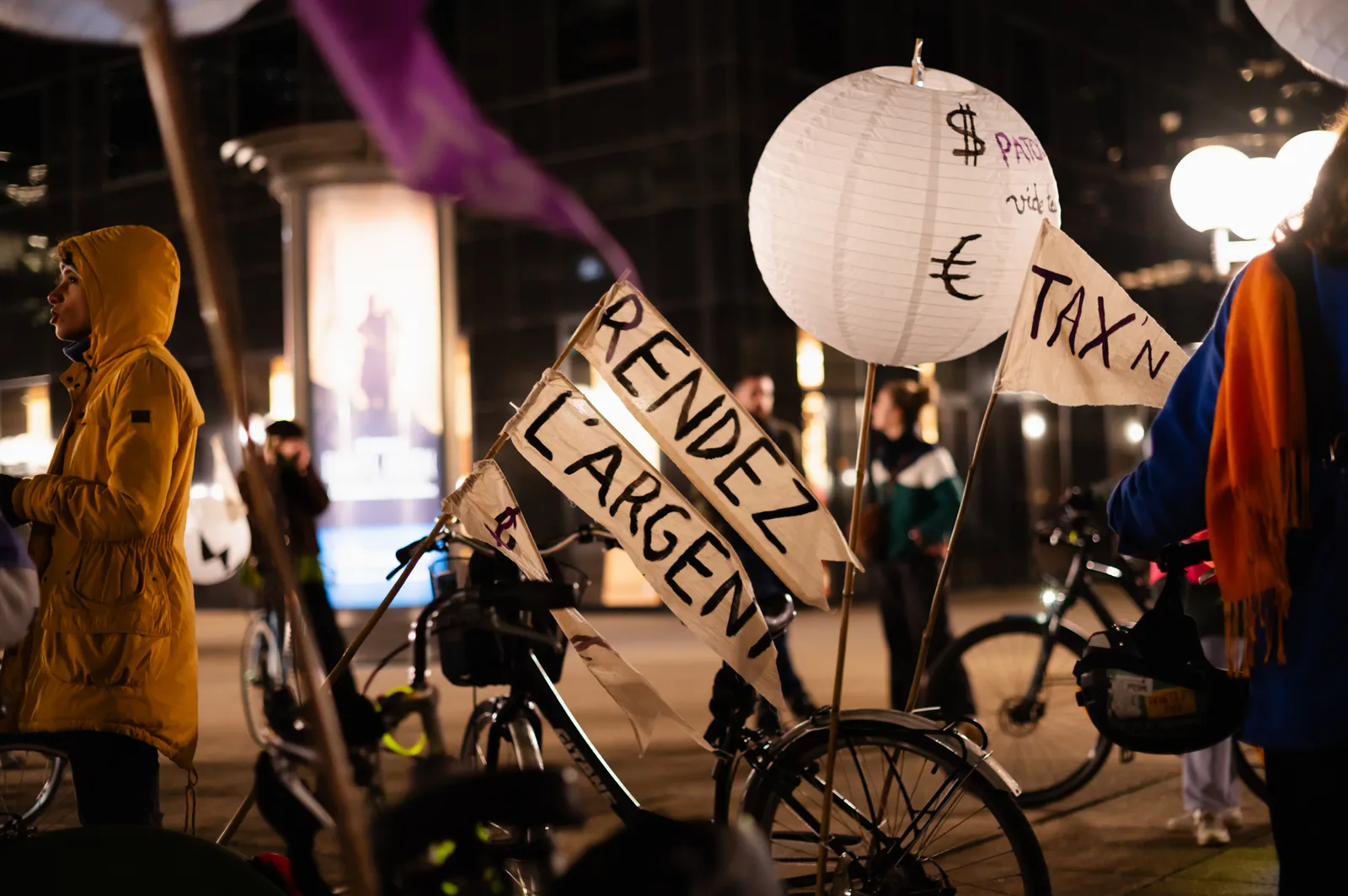 A nighttime protest scene with cyclists holding signs reading "RENDEZ L'ARGENT" and a paper lantern marked with currency symbols including dollar and euro signs. Protesters wear colorful rain gear.