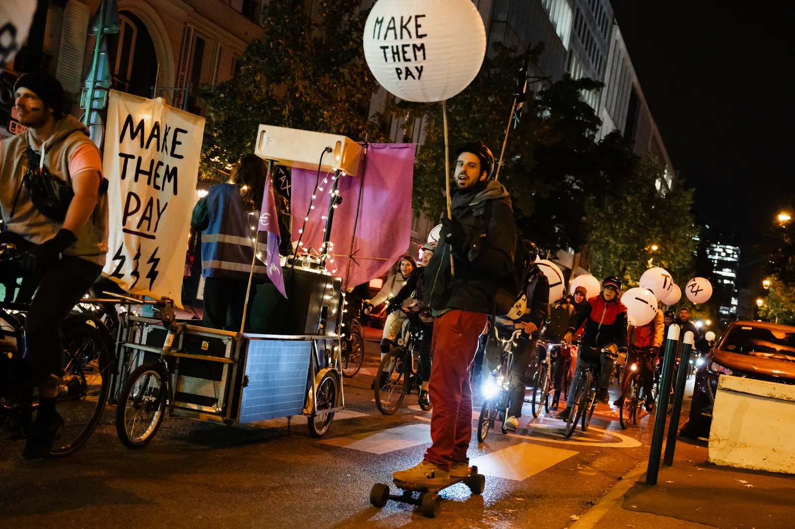 Night protest with cyclists holding "MAKE THEM PAY" signs, featuring illuminated banners and creative mobile displays on bikes gathering in an urban street demonstration.