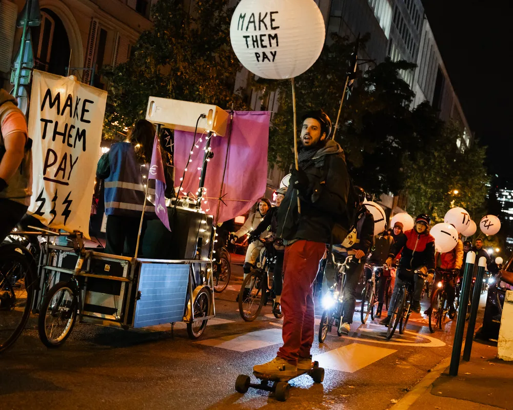 Night protest with cyclists holding "MAKE THEM PAY" signs, featuring illuminated banners and creative mobile displays on bikes gathering in an urban street demonstration.