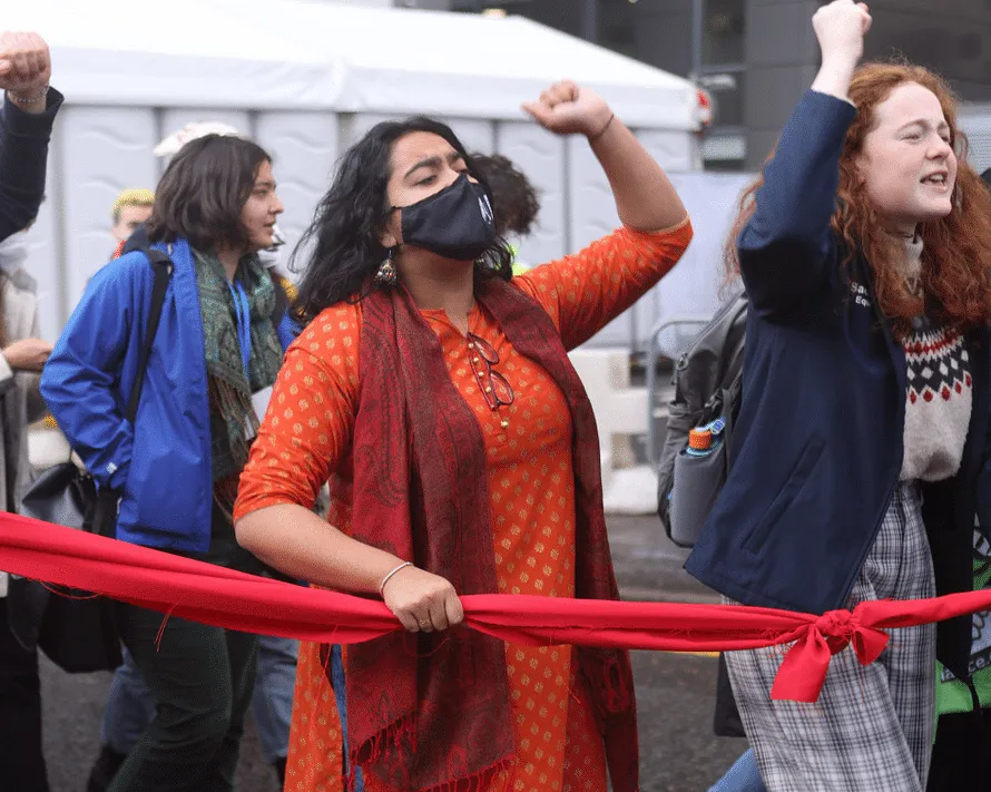 A woman in an orange sari is holding a red line, fist raised, during the final day of COP26