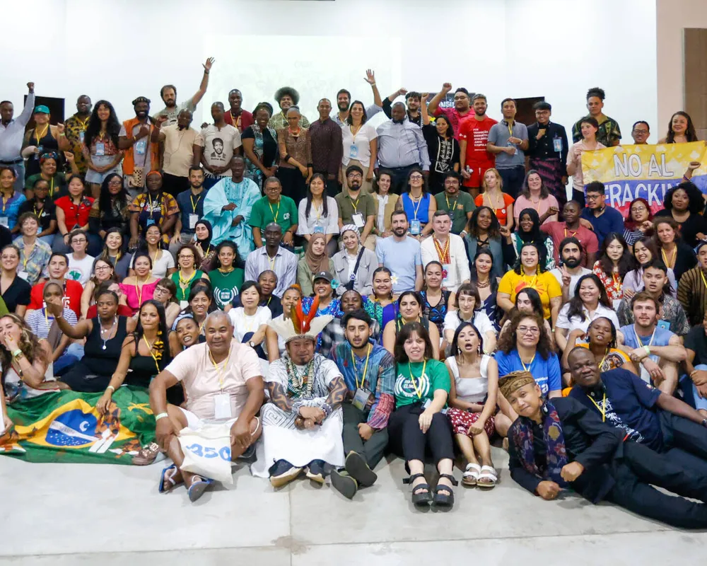 Large group photo of over 100 global activists gathered indoors, at an activist gathering event, displaying flags and banners showing where they come from.