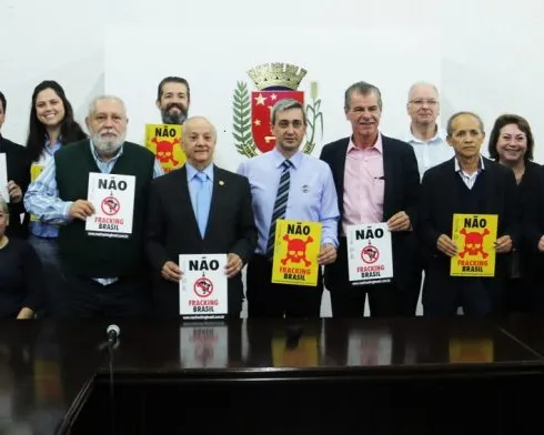 A group of people standing behind a conference table holding protest signs with "NAO" (No) written on them, appearing to be at a formal meeting or press conference opposing some initiative.